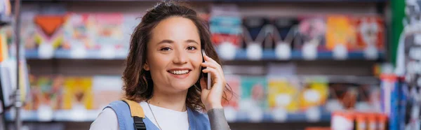 Happy brunette woman looking at camera and talking on cellphone in blurred stationery shop, banner — Photo de stock