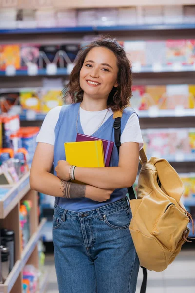 Татуйована жінка з новими блокнотами і рюкзаком посміхається на камеру в канцелярському магазині — стокове фото