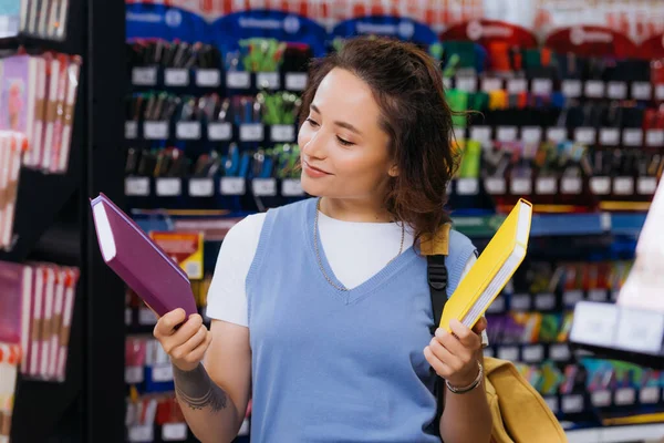 Lächelnder tätowierter Student, der in verschwommenem Schreibwarenladen Werbebücher auswählt — Stockfoto