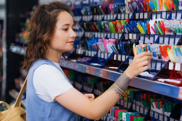 Young tattooed woman near rack with multicolored pens in stationery store - foto de stock