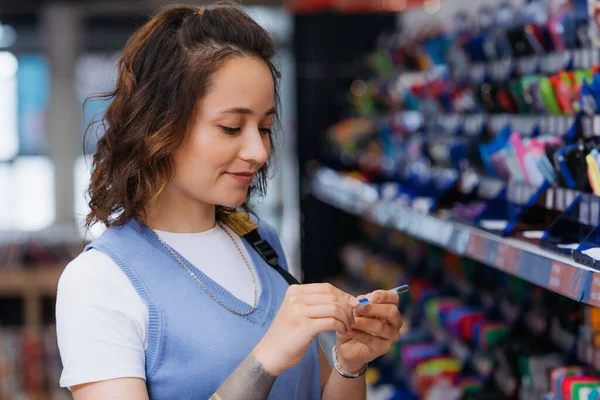 Позитивна брюнетка жінка з ручкою в розмитому канцелярському магазині — стокове фото