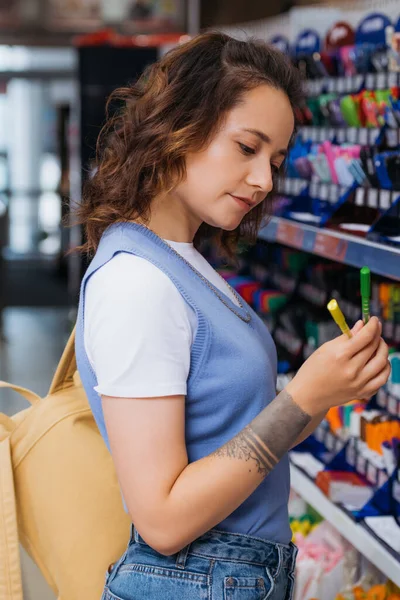 Tattooed student with backpack holding colorful pens in stationery shop - foto de stock