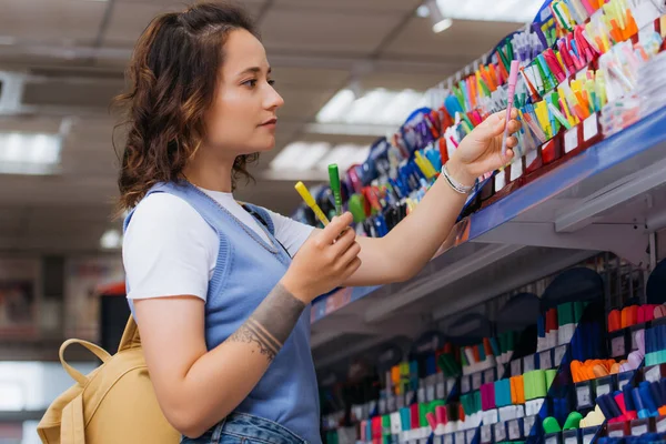 Side view of tattooed woman holding multicolored pens near rack in shop — Stock Photo