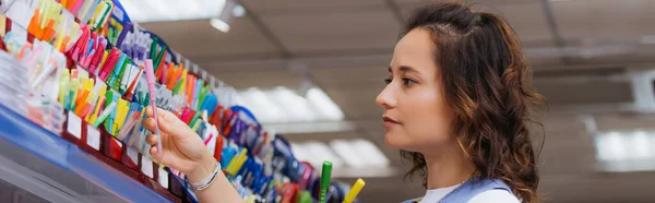 Junge brünette Frau wählt Stift auf Regal in Schreibwarengeschäft, Banner — Stockfoto