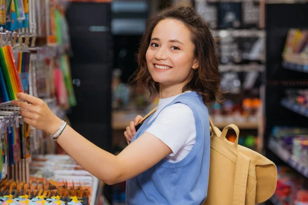 Pretty woman with backpack smiling at camera while choosing felt pens in store — Fotografia de Stock