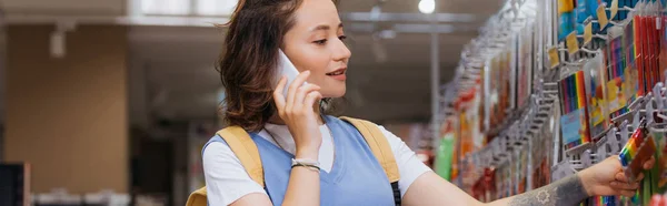 Brunette woman choosing felt pens while talking on smartphone in stationery store, banner — Stock Photo