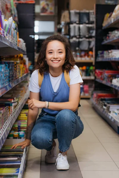 Весела молода жінка в джинсах дивиться на камеру біля стелажів в канцелярському магазині — стокове фото