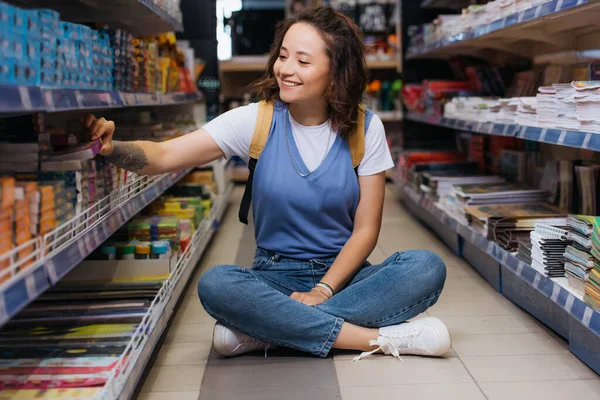 Glückliche Frau sitzt mit gekreuzten Beinen zwischen Regalen im Schreibwarengeschäft — Stockfoto