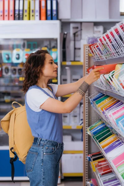Side view of student with backpack choosing copybook on rack with stationery — стоковое фото