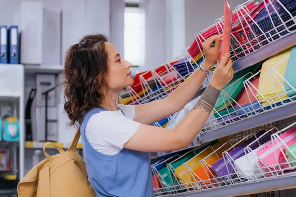 Side view of tattooed student with backpack choosing plastic folder in stationery store — стоковое фото