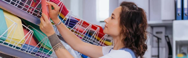Tattooed woman near rack with colorful plastic folders in stationery shop, banner — Stock Photo