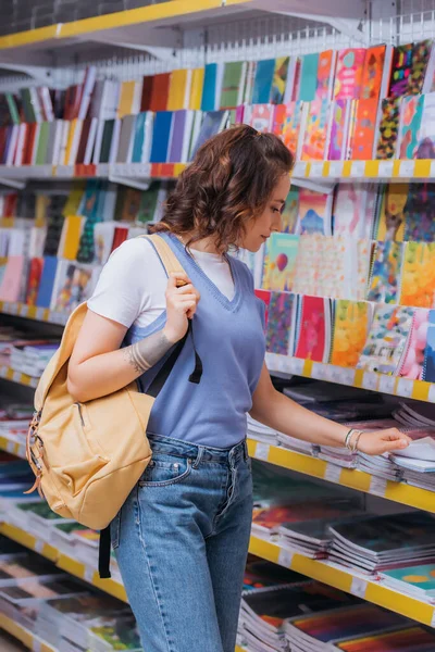 Studentin mit Rucksack neben Regal mit bunten Heften im Schreibwarengeschäft — Stockfoto