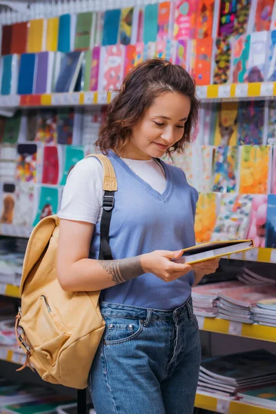 Pleased brunette woman with backpack holding new notebook in blurred store — Stockfoto