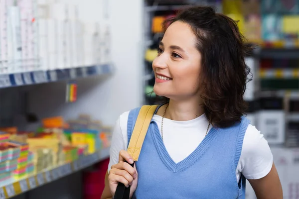 Happy young woman with backpack near blurred rack with stationery — Stock Photo