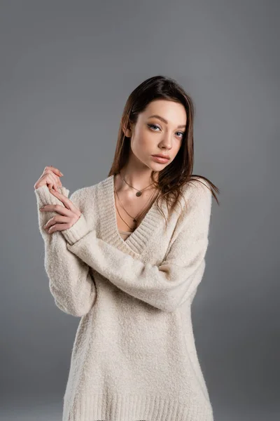 Young woman in cozy sweater and golden necklaces looking at camera isolated on grey — Stockfoto