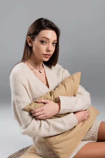 Young woman in sweater and golden necklaces hugging pillow on grey background — Stock Photo