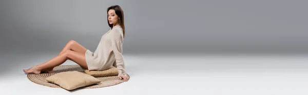 Full length of woman with slim naked legs sitting on carpet in soft sweater on grey background, banner — стоковое фото