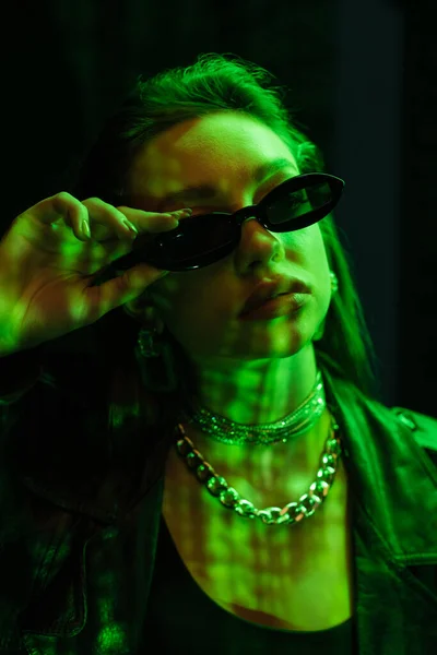 Portrait of trendy woman in silver accessories adjusting sunglasses in green neon light isolated on black — Stock Photo
