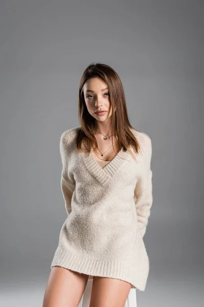 Young woman in cozy sweater standing with hands behind back and looking at camera on grey background — стоковое фото