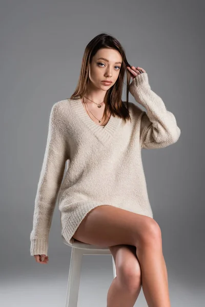Young woman in long sweater touching hair and looking at camera while sitting on grey background — Photo de stock