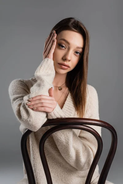 Brunette woman in soft sweater sitting on chair and touching face isolated on grey - foto de stock