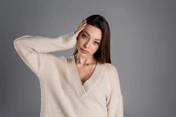 Young brunette woman in soft sweater touching head and looking at camera isolated on grey - foto de stock