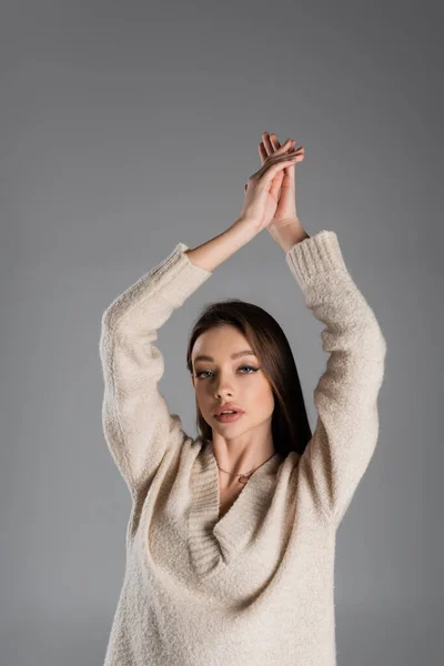 Чуттєва жінка в теплому светрі позує з піднятими руками ізольовано на сірому — стокове фото