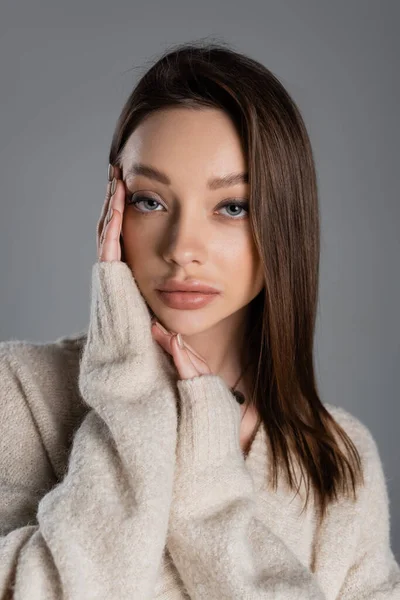 Brunette woman with makeup holding hands near face and looking at camera isolated on grey — стоковое фото