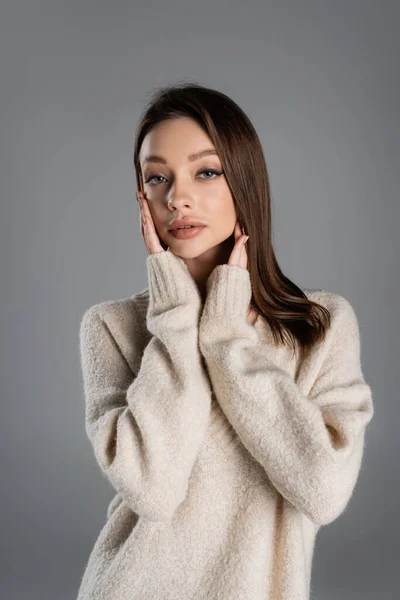Pretty woman in cozy sweater touching face while looking at camera isolated on grey — Stockfoto