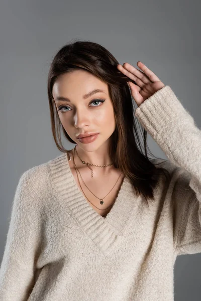 Young woman in sweater and golden necklaces touching hair isolated on grey — Stockfoto