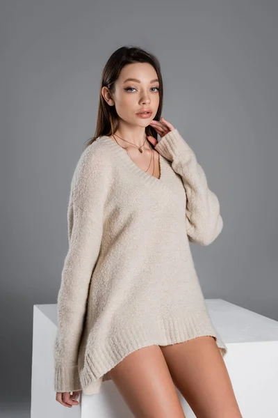 Young woman in soft sweater and golden necklaces sitting on white cube isolated on grey — Stockfoto
