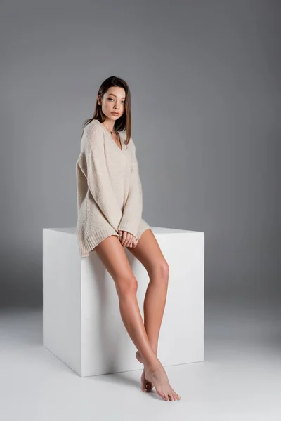 Full length of woman with slender naked legs sitting in sweater on white cube on grey background — Stock Photo