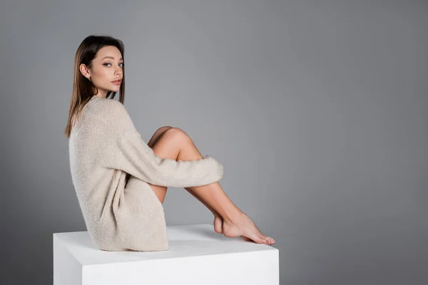 Повна довжина жінки в затишному светрі, обіймаючи голі ноги, сидячи на білому кубі ізольовані на сірому — стокове фото