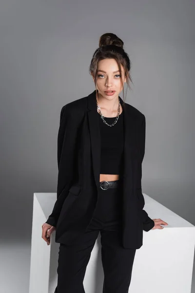 Brunette woman in black suit and silver necklace looking at camera near white cube on grey background — Photo de stock