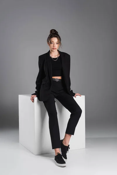 Full length of woman in black sneakers and suit sitting on white cube on grey background — Stockfoto