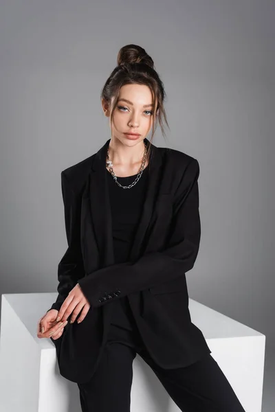 Trendy woman in black jacket sitting on white cube and looking at camera isolated on grey — Stockfoto