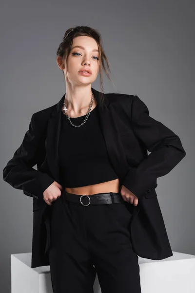 Young and stylish woman in black clothes and metal necklaces standing with hands on hips isolated on grey - foto de stock