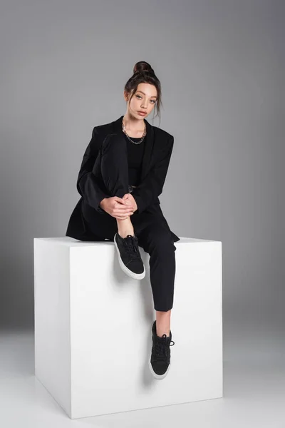 Full length of woman in black suit and sneakers sitting on white cube on grey background — стоковое фото