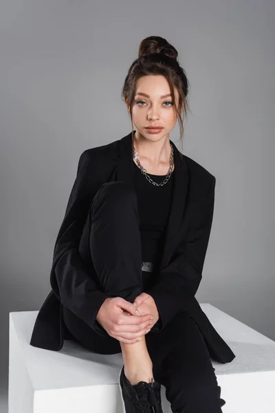 Charming woman in black jacket sitting on white cube and looking at camera isolated on grey — стоковое фото