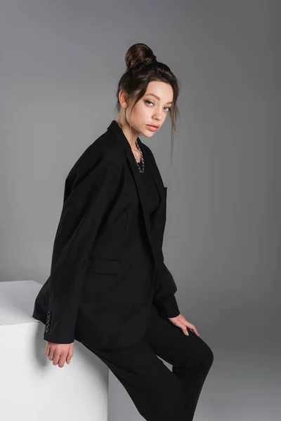 Young and trendy woman in black blazer looking at camera near white cube isolated on grey — Stock Photo