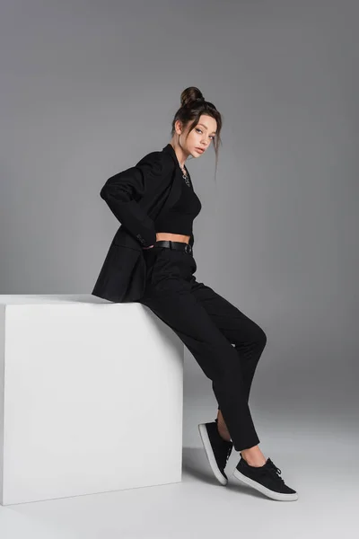 Full length of woman in black trendy clothing sitting on white cube with hand on hip on grey background — Photo de stock