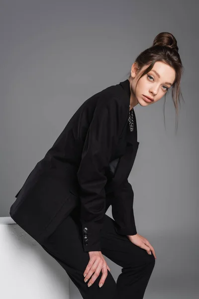 Brunette woman in black suit sitting on white cube and looking at camera isolated on grey — Stockfoto