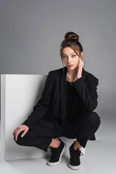 Fashionable woman in black suit sitting on haunches near white cube on grey background — Foto stock