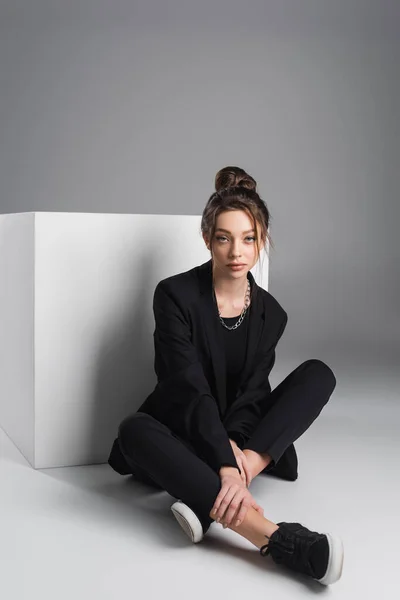 Stylish woman in black suit and sneakers sitting with crossed legs near white cube on grey background - foto de stock