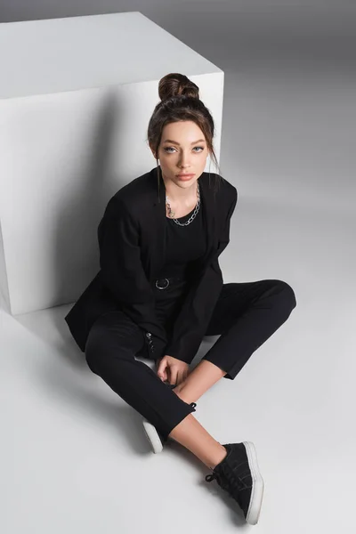 Full length of woman in black suit sitting with crossed legs near white cube on grey background — Fotografia de Stock