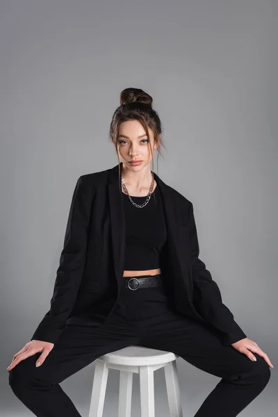 Stylish brunette woman in black suit and metal necklace sitting and looking at camera isolated on grey — Stockfoto