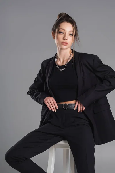 Sensual woman in black suit and silver necklace sitting with hands on waist isolated on grey — стоковое фото