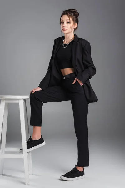 Young brunette woman posing with hand in pocket of black trousers near white stool on grey background — Stock Photo