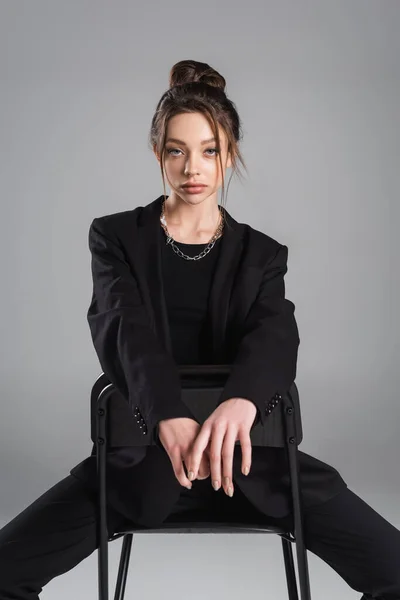 Brunette woman in black clothes sitting on chair and looking at camera isolated on grey — стоковое фото