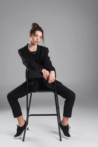 Full length of young woman in black suit and sneakers sitting on chair on grey background — Stockfoto